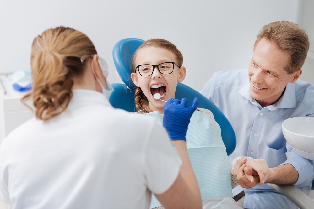 Young girl with braces receiving a dental exam at a family dentistry practice, with her father seated beside her for support
