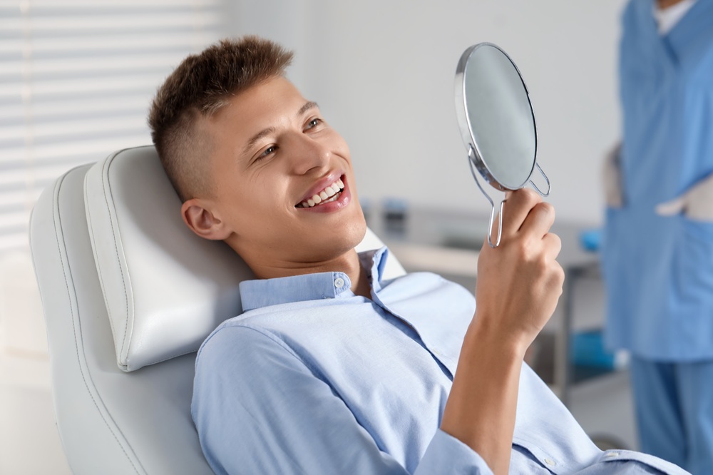 Young male patient sitting in a dental chair, smiling and holding a mirror to admire his results after cosmetic dental treatment at Prescott Dental Arts in Prescott, AZ