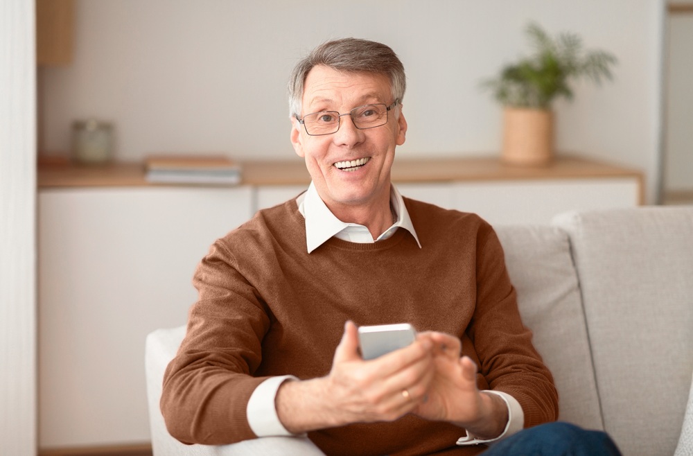Affordable Dental Implants, Dentist Prescott AZ Older man smiling confidently at home while holding a phone, representing the peace of mind that comes with affordable dental implant options at Prescott Dental Arts in Prescott, AZ